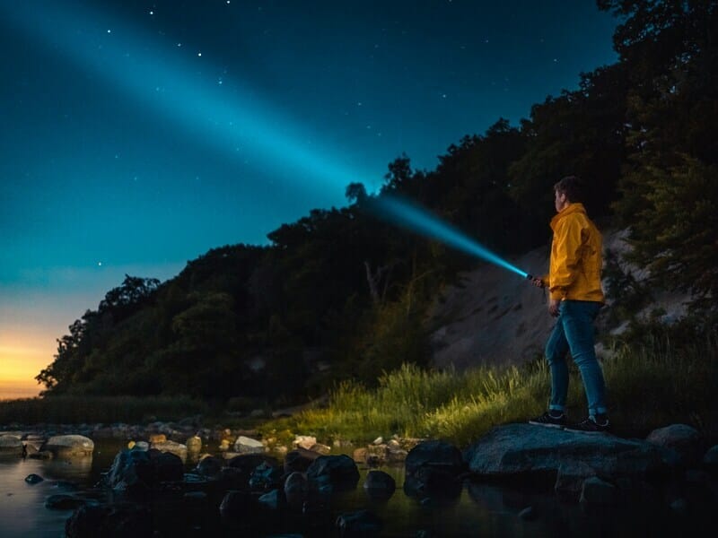 A man in a yellow jacket shining a flashlight into the night sky.